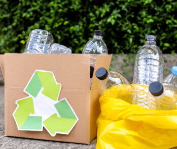 Garrafas plásticas descartadas em caixa de papelão com símbolo de reciclagem e saco amarelo, representando a aplicação da política dos 5Rs na gestão de resíduos.