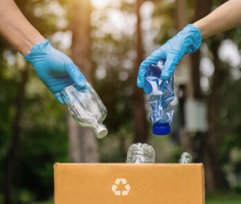 Mãos com luvas descartando garrafa de vidro e plástico em caixa de coleta seletiva com símbolo de reciclagem ao ar livre