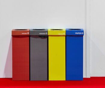 Lixeiras coloridas para coleta seletiva: plástico (vermelha), rejeitos (cinza), metal (amarela) e papel (azul), em ambiente interno com piso vermelho.
