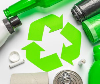 Símbolo da reciclagem cercado por materiais recicláveis como plástico, vidro, papelão, alumínio e sacos de lixo, representando a coleta seletiva.