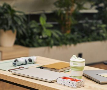 Mesa de escritório sustentável com copo reutilizável, cadernos ecológicos e vegetação ao fundo, representando ações sustentáveis na gestão corporativa de resíduos.