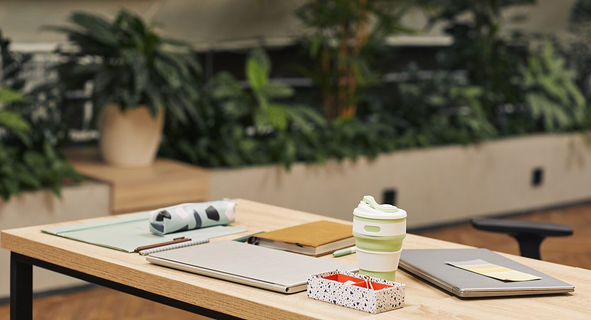 Mesa de escritório sustentável com copo reutilizável, cadernos ecológicos e vegetação ao fundo, representando ações sustentáveis na gestão corporativa de resíduos.