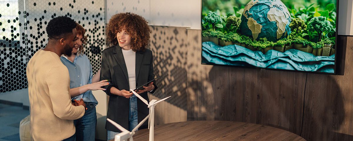 Profissionais discutem sustentabilidade corporativa em sala com maquete de turbinas eólica e gráfico ESG, ao lado de tela com imagem do planeta Terra