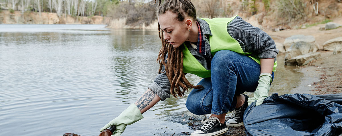 Jovem com dreadlocks, vestindo colete de voluntariado verde limão e luvas, realiza a limpeza da margem de um lago ou rio, retirando uma garrafa plástica da água. Um saco de lixo preto está ao seu lado, indicando a coleta de resíduos para diminuir o impacto ambiental.