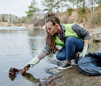 Jovem com dreadlocks, vestindo colete de voluntariado verde limão e luvas, realiza a limpeza da margem de um lago ou rio, retirando uma garrafa plástica da água. Um saco de lixo preto está ao seu lado, indicando a coleta de resíduos para diminuir o impacto ambiental.