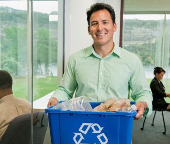 Homem segurando caixa azul de reciclagem com resíduos plásticos em escritório, representando ações práticas para redução da geração de resíduos na empresa.