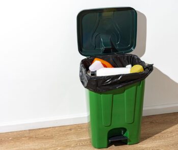 Lixeira verde com saco plástico preto contendo resíduos recicláveis, como embalagens plásticas, representando a separação correta de resíduos sólidos urbanos.