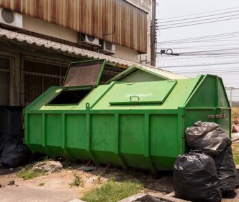 Caçamba de entulho verde com sacos de lixo ao redor, posicionada nos fundos de um estabelecimento comercial.