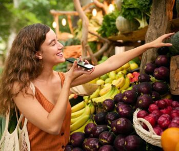 Mulher praticando o consumo consciente ao comprar produtos frescos em um mercado, carregando uma sacola retornável.