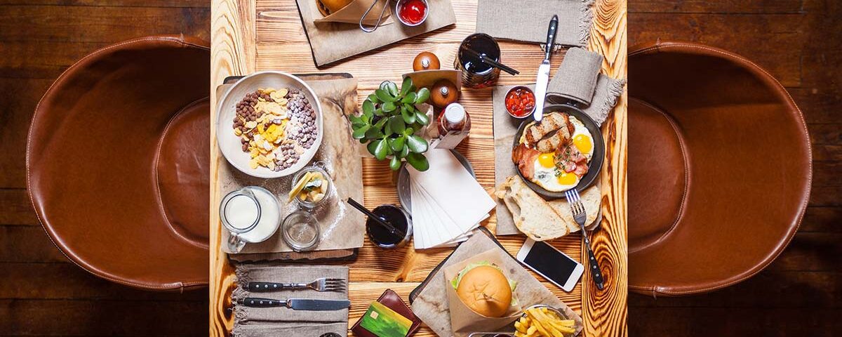 Mesa de restaurante vista de cima com hambúrgueres, batatas fritas, ovos com bacon, cereais e bebidas, ilustrando o consumo de alimentos e a geração de resíduos orgânicos e recicláveis no setor de food service.