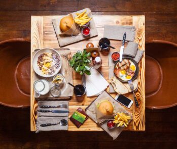 Mesa de restaurante vista de cima com hambúrgueres, batatas fritas, ovos com bacon, cereais e bebidas, ilustrando o consumo de alimentos e a geração de resíduos orgânicos e recicláveis no setor de food service.