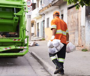 Coletor de lixo realizando coleta de resíduos em área urbana, próximo a caminhão de lixo verde. Conceitos de gestão de resíduos, coleta seletiva e diferença entre lixo, resíduo e rejeito.
