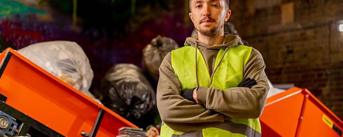 Homem com colete de segurança em central de triagem de resíduos, com lixo e equipamentos visíveis ao redor.