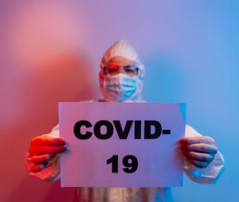 Profissional da saúde com equipamento de proteção individual (EPI) segurando cartaz com a palavra COVID-19, simbolizando os impactos ambientais da pandemia.