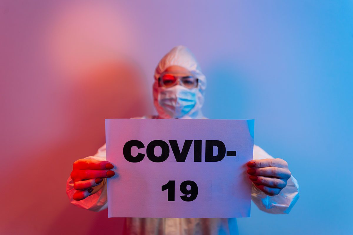 Profissional da saúde com equipamento de proteção individual (EPI) segurando cartaz com a palavra COVID-19, simbolizando os impactos ambientais da pandemia.