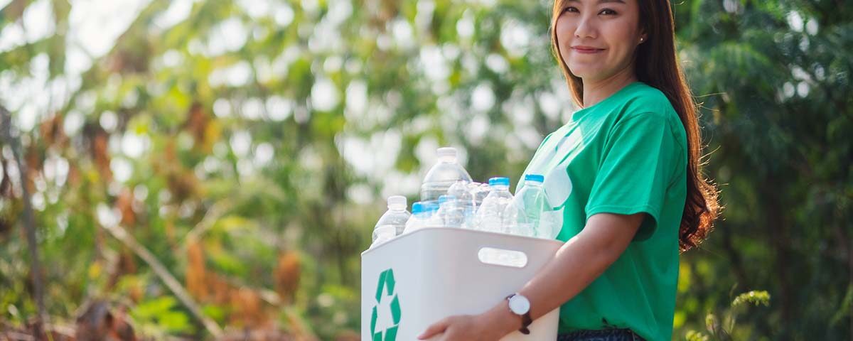 Mulher de camiseta verde segura uma caixa com o símbolo da reciclagem cheia de garrafas PET, em um parque.
