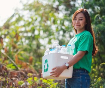 Mulher de camiseta verde segura uma caixa com o símbolo da reciclagem cheia de garrafas PET, em um parque.