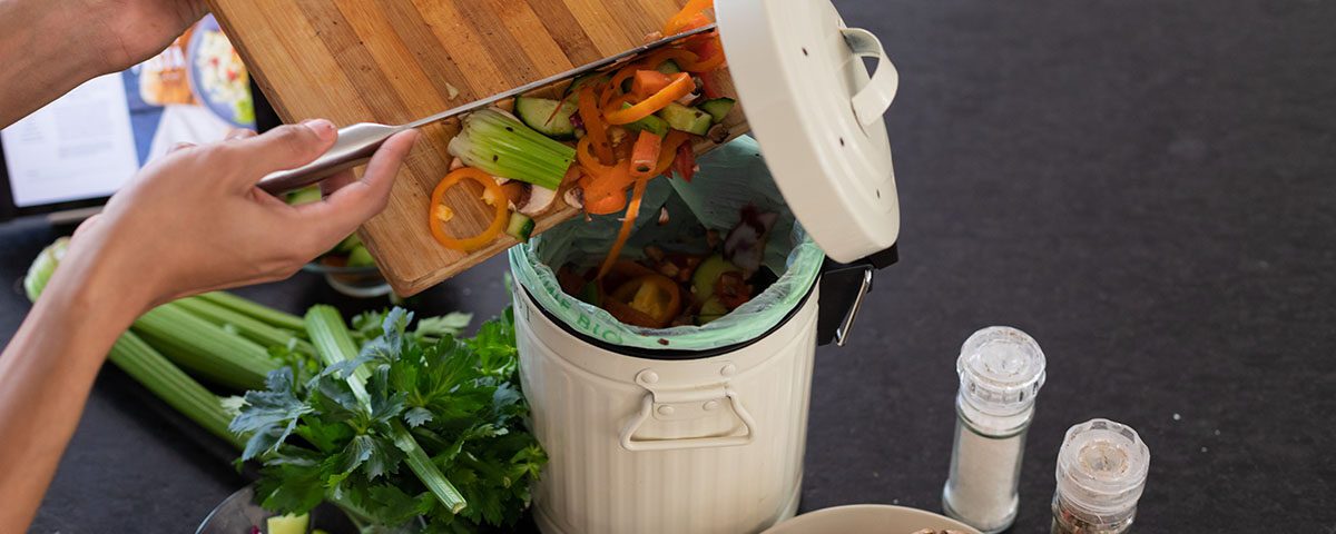 Pessoa descartando restos de vegetais crus em uma lixeira de compostagem doméstica, ao lado de alimentos frescos, representando o reaproveitamento de resíduos orgânicos na compostagem.