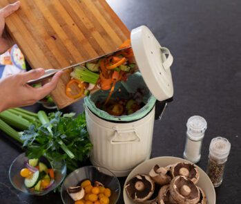 Pessoa descartando restos de vegetais crus em uma lixeira de compostagem doméstica, ao lado de alimentos frescos, representando o reaproveitamento de resíduos orgânicos na compostagem.