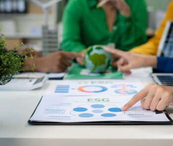 Equipe discutindo estratégias ESG (Environmental, Social and Governance) em escritório sustentável, com gráficos, relatórios de impacto ambiental e energia renovável representada por painel solar e globo terrestre.