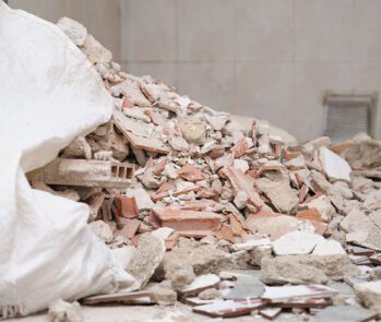 Imagem ilustrativa de resíduos da construção civil, composta por entulho de tijolos, concreto, argamassa e cerâmica, dispostos em um ambiente interno. A cena representa práticas comuns de descarte e gestão de resíduos sólidos na construção, fundamentais para a sustentabilidade e o cumprimento da legislação ambiental brasileira. Ideal para conteúdos sobre reciclagem de entulho, gestão de resíduos da construção civil (RCC), logística reversa e sustentabilidade no setor da construção.