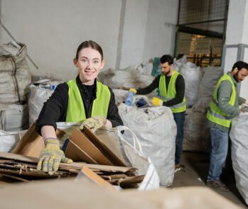 Trabalhadores com colete de segurança realizando triagem de resíduos recicláveis em centro de gestão de resíduos, seguindo normas de saúde e segurança do trabalho.