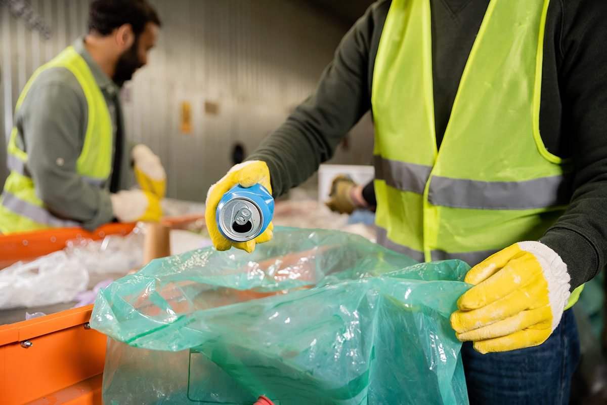 Profissional com colete de segurança manuseando lata de alumínio em centro de triagem de sucata, demonstrando práticas corretas de destinação de resíduos recicláveis conforme a legislação ambiental.